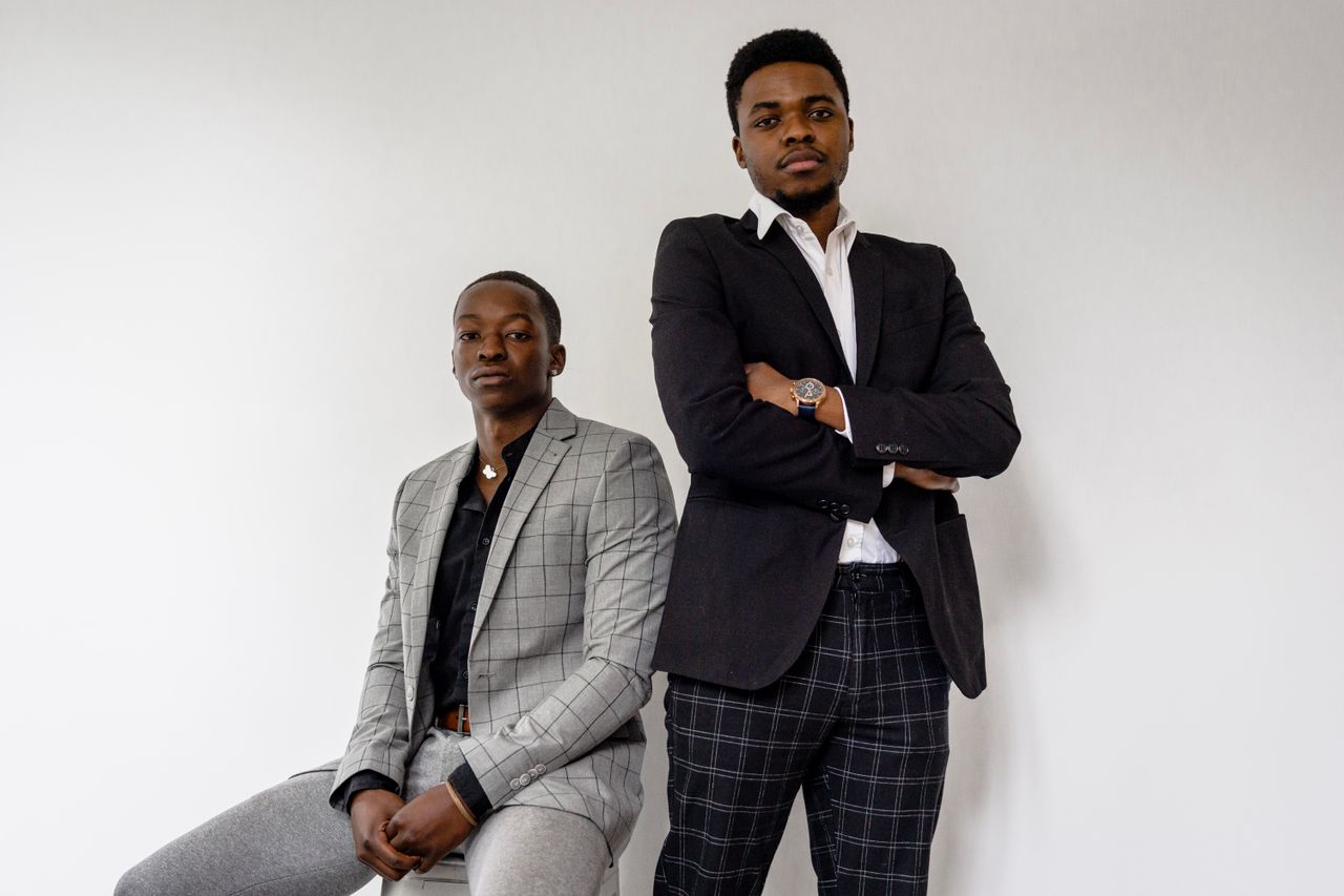 Portrait of two men in suits, one sitting, one standing, on a white background.